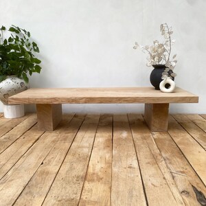 May include: A natural wood coffee table with a rectangular top and two block-style legs. The table is made of light-colored wood and sits on a wooden floor. A black vase and a white vase are on the table.
