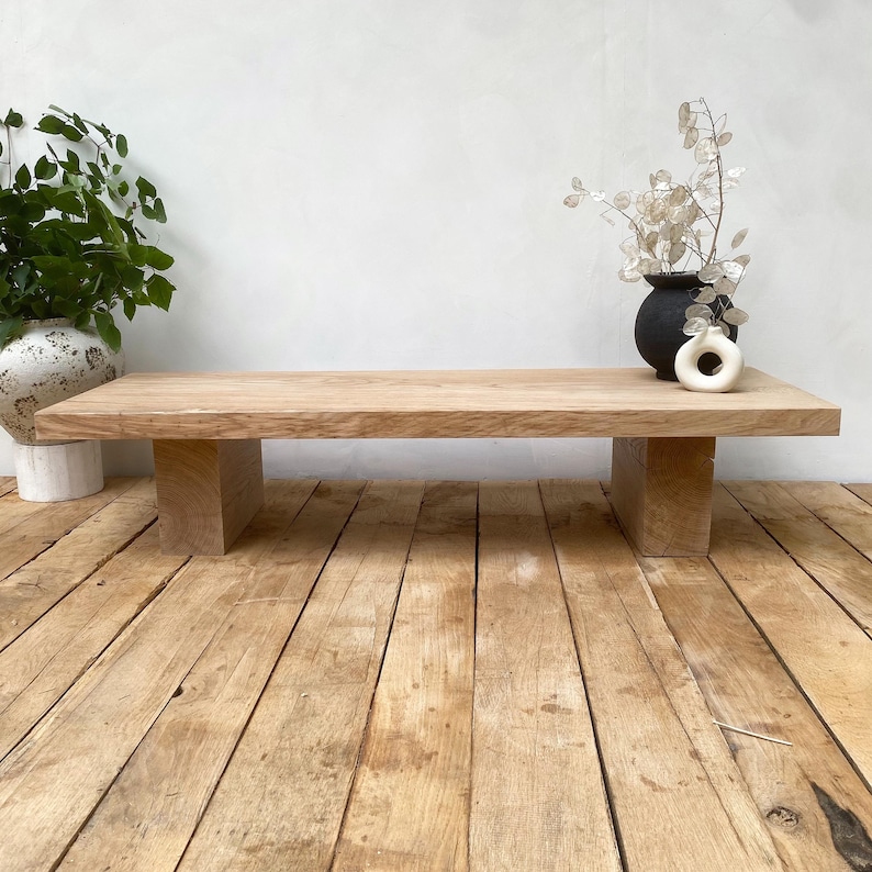 May include: A natural wood coffee table with a rectangular top and two block-style legs. The table is made of light-colored wood and sits on a wooden floor. A black vase and a white vase are on the table.