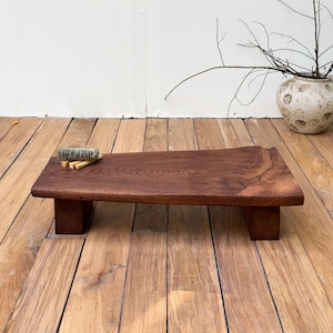 May include: A low rectangular wooden table with a dark brown finish and a natural live edge on one side. The table features a visible wood grain pattern and is supported by two block legs. A small bundle of dried sage rests on the surface.