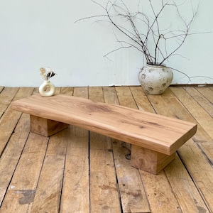 May include: A low rectangular wooden bench featuring a natural light brown wood grain finish with a smooth top surface and two sturdy block-style legs. The bench is crafted from solid wood with visible knots and organic textures on the edges.
