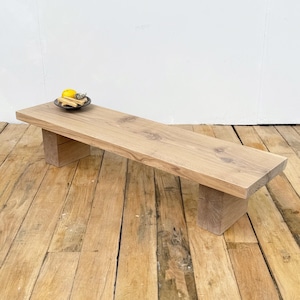 May include: A light-coloured wooden bench with a rectangular top and two block-style legs. A small, dark-coloured dish with a lemon and other items sits on the bench. The bench is made of natural wood and has a rustic appearance.