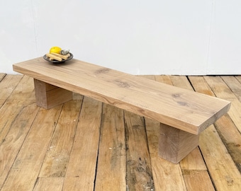 May include: A light-colored wooden bench with a rectangular top and two block-style legs. A small, dark-colored dish with a lemon and other items sits on the bench. The bench is made of natural wood and has a rustic appearance.