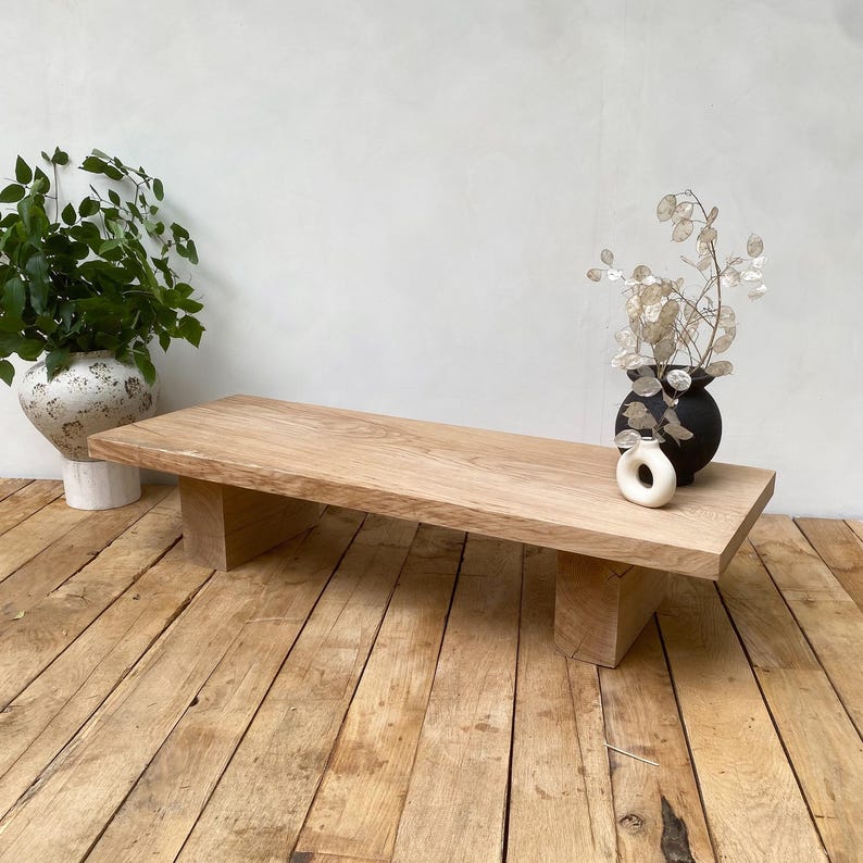 May include: A light-colored, rectangular wooden coffee table with a minimalist design. The table has a thick, solid top supported by two rectangular legs. The table is placed on a wooden floor. A vase with greenery and a black vase with dried flowers are on the table.