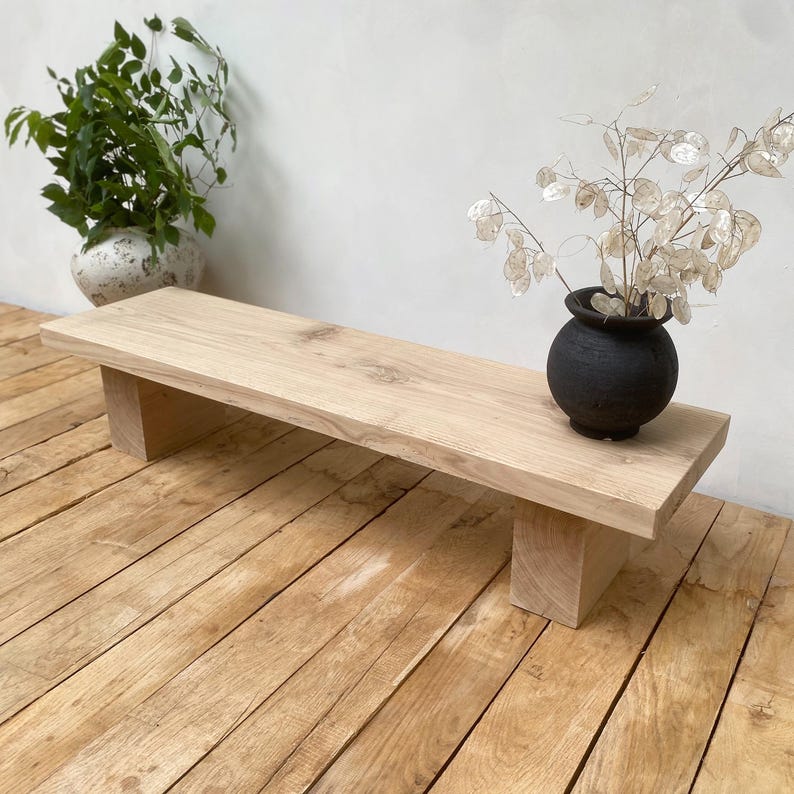 May include: A light-colored wooden bench with a rectangular top and two supporting blocks. A small black vase with dried flowers sits on the bench. The bench is on a wooden floor, and a white vase with greenery is in the background.