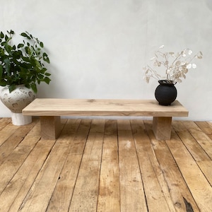 May include: A light-colored wooden bench with a rectangular top and two block-shaped legs. The bench is placed on a wooden floor. A white and silver vase with green leaves and a black vase with dried flowers are on the bench.