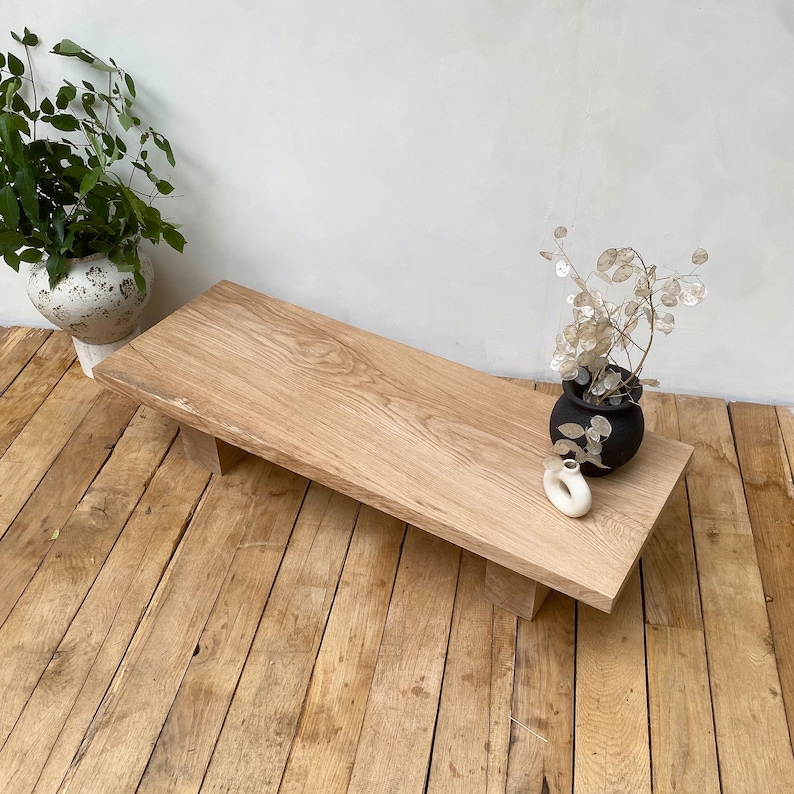 May include: A rectangular, light-colored wooden coffee table with a natural wood grain finish. The table is supported by two short, rectangular legs. A small black vase with dried flowers and a white ceramic object sit on the table.