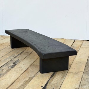 May include: A black wooden bench with a curved seat and two rectangular legs. The bench is made of solid wood and has a natural, textured finish. The bench is approximately 48 inches long.