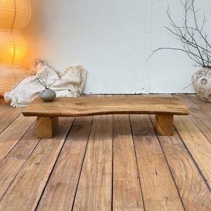 May include: A low rectangular wooden bench featuring a natural live edge top and two thick block legs. The bench is crafted from light brown wood with a visible grain pattern, showcasing a smooth surface and a sturdy, minimalist construction.
