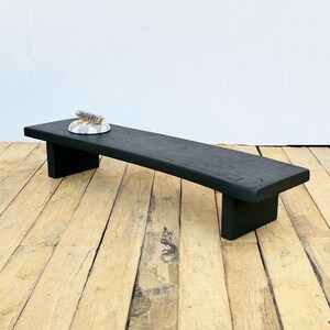May include: A black wooden bench with a rectangular top and two rectangular legs. A small, round, marble-patterned tray with a bundle of sage is on the bench. The bench is approximately 48 inches long.