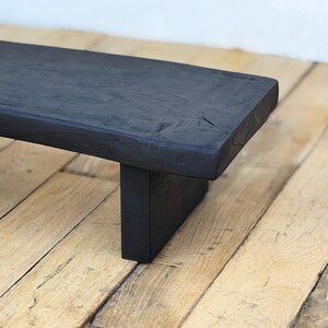 May include: A black wooden bench with a rectangular top and a single, centered leg. The bench is made of dark-stained wood, and the surface shows the natural grain and texture of the wood. The bench is approximately 36 inches long.