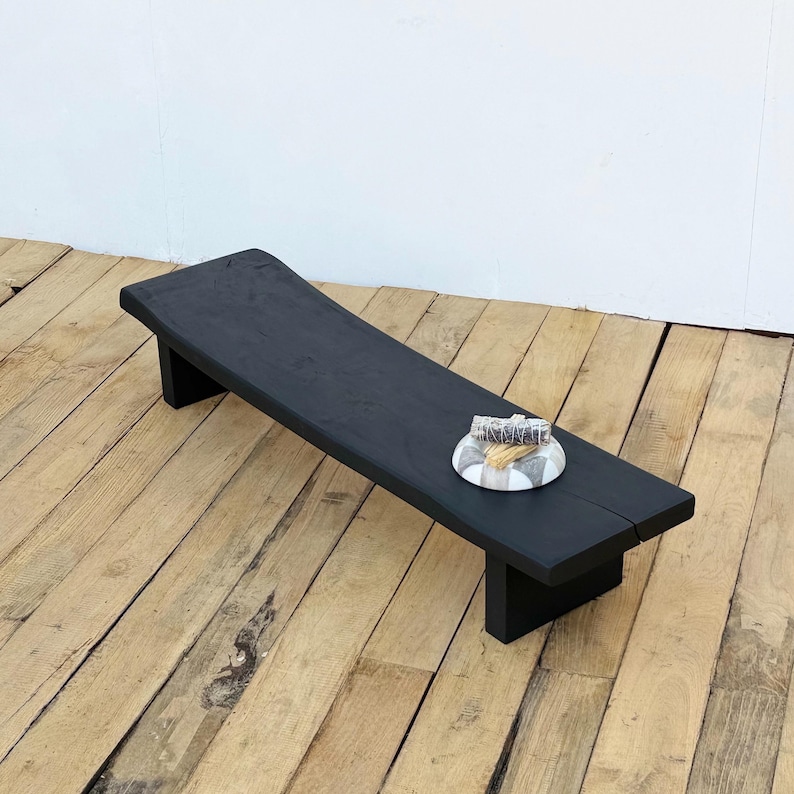 May include: A black wooden bench with a rectangular top and two short legs. The bench has a natural, slightly curved edge. A small, round decorative plate with a gray and white pattern sits on the bench.