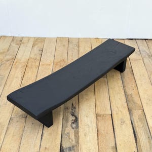 May include: A black, rectangular wooden riser with a slightly curved top. The riser is supported by two short legs, one at each end. The wood grain is visible, and the finish is matte. The riser is designed to elevate items.