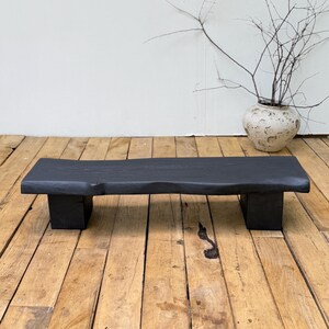 Peut inclure: Un banc bas rectangulaire en bois massif avec une finition noire mate. Le banc présente une surface supérieure épaisse à bord naturel, soutenue par deux pieds robustes en forme de bloc, révélant une texture de grain de bois visible.