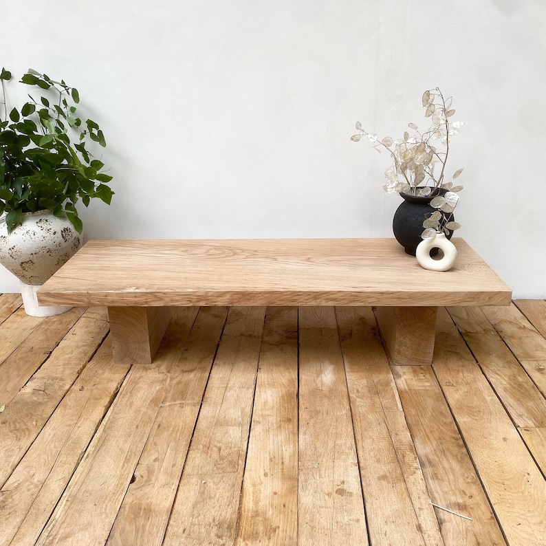 May include: A light-colored, rectangular wooden coffee table with a natural finish. The table is supported by two thick, rectangular legs. A black vase with dried flowers and a white ceramic ring are on the table. A potted plant is on the left.