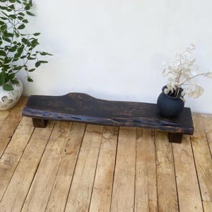 Può includere: Un vassoio da portata in legno marrone scuro e rustico con un bordo naturale, appoggiato su due brevi supporti in legno marrone scuro. Un piccolo vaso di ceramica nera con fiori secchi è posto sul vassoio. Il vassoio è esposto su un pavimento in legno marrone chiaro.