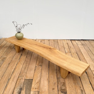 May include: A light-coloured wooden bench with a natural finish. The bench is rectangular with a live edge and stands on four short, angled legs. A small, round, green vase with dried branches sits on the bench.