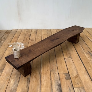 May include: A long, dark brown wooden bench with a natural live edge top and two thick, block-style legs. A small, cylindrical, light-colored stone or ceramic vase containing dried, translucent, circular plant stems sits on the left side.