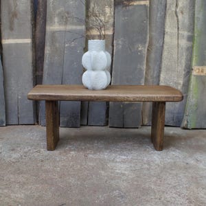 May include: A small, rectangular wooden coffee table with rounded corners and four legs. A white, textured vase with a distinctive, rounded design is placed on the table, containing thin, dark branches. The background is made of weathered wooden planks.