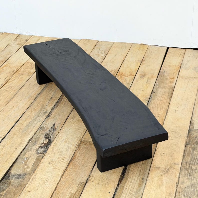 May include: A black, curved wooden bench with a rectangular top and two short, rectangular legs. The bench is made of solid wood and has a matte finish. The bench is designed for indoor or outdoor use.
