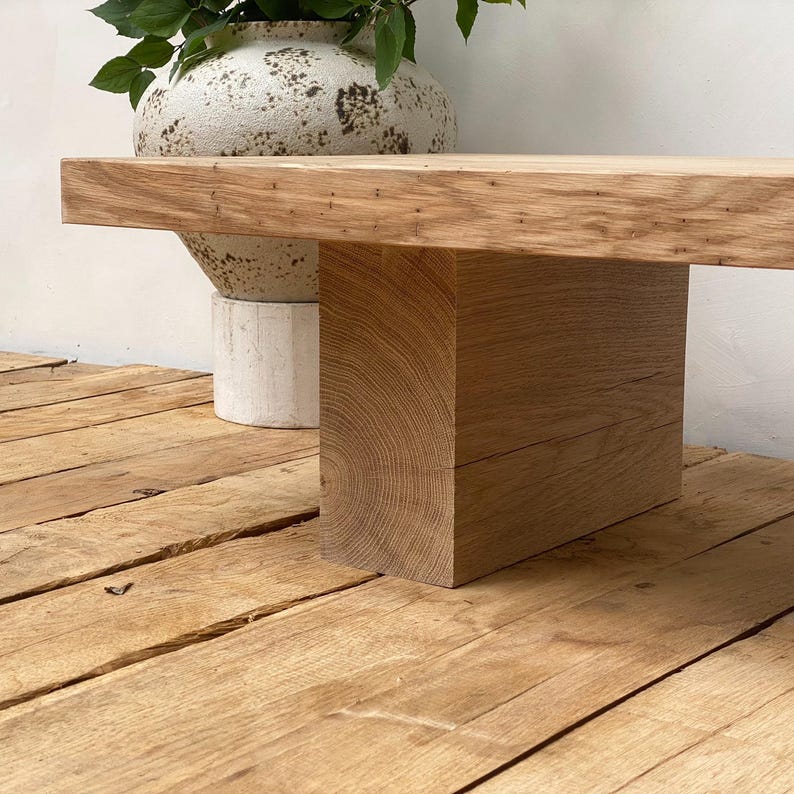 May include: A solid oak wood bench with a rectangular top and a thick, square base. The bench is light brown and sits on a wooden floor. A decorative vase with green leaves is in the background.
