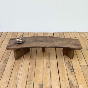 May include: A handcrafted, solid wood coffee table with a natural, live edge design. The table is stained a warm brown and features a low profile with sturdy, angled legs. A small, round dish with wooden pieces sits on the table's surface.