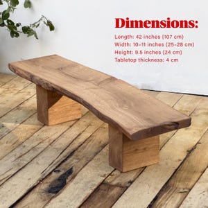 May include: A wooden bench with a live edge design. The bench has two rectangular wooden legs. Dimensions: 107 cm long, 25-28 cm wide, and 24 cm high. Tabletop thickness: 4 cm. The bench is made of natural wood.