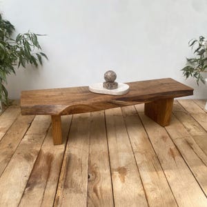 May include: A rustic wooden bench with a natural edge and two sturdy legs. A small, oval, white marble tray holds a decorative stone sphere and a smaller, stacked stone sculpture. The bench is made of light brown wood.
