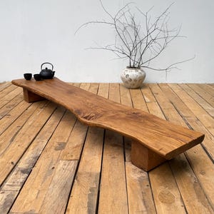 May include: A long, low-profile wooden bench featuring a natural live edge and a warm brown wood grain finish. The bench is supported by two thick, rectangular wooden block legs and is topped with a small black teapot and two matching cups.