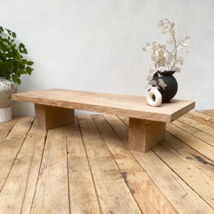 Peut inclure: Une table basse rectangulaire en bois clair avec deux pieds en forme de blocs. Un vase noir avec des fleurs séchées et un petit objet blanc en forme d'anneau sont posés sur la table. La table est sur un plancher en bois.