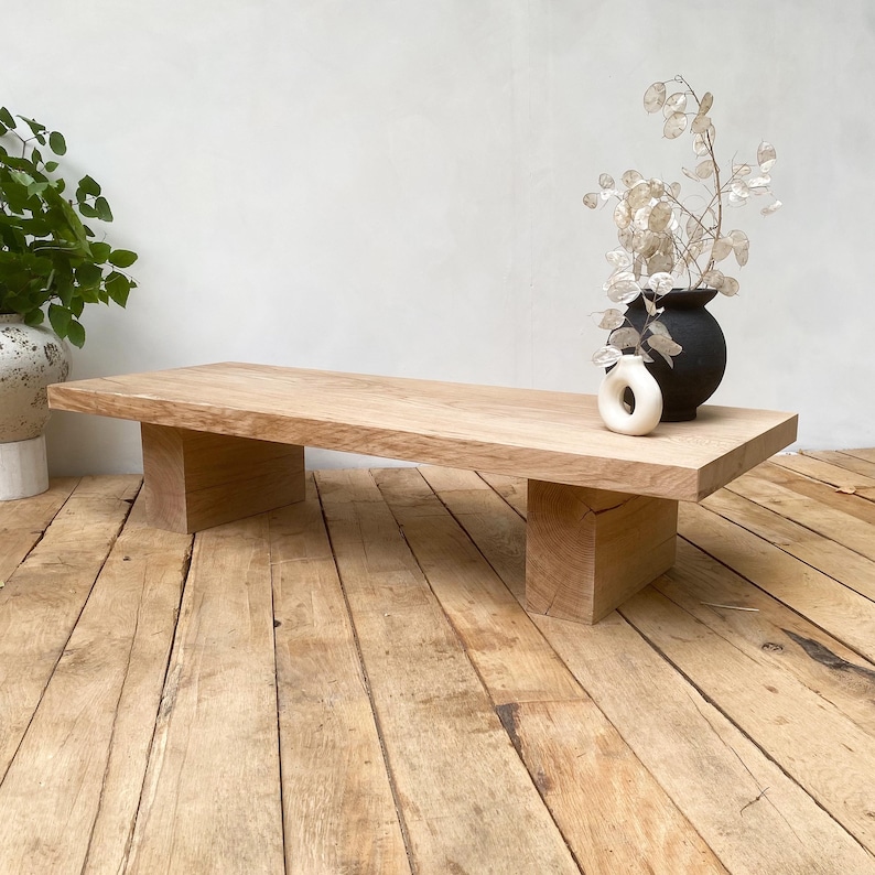 May include: A light-colored, rectangular wooden coffee table with two block-style legs. A black vase with dried flowers and a small white ring-shaped object sit on the table. The table is on a wooden floor.