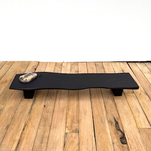 May include: A black wooden serving tray with a distinctive, wavy edge. The tray is supported by two short, rectangular legs. A small, decorative dish with a patterned design sits on the tray's surface.