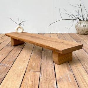 May include: A rectangular wooden riser with a natural light brown wood grain finish, supported by two thick, semi-circular block legs. The surface is flat and smooth, showcasing the organic texture and warm tones of the wood material.