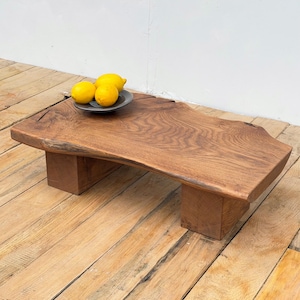 May include: A rustic, wooden coffee table with a live edge design. The table is supported by two rectangular legs. Three yellow lemons sit on a small, grey plate on the table's surface. The table is made of solid wood, approximately 120cm long.