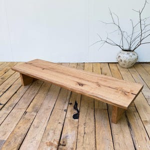 Peut inclure: Un banc bas rectangulaire fabriqué en bois naturel brun clair avec un grain visible. Le banc présente une surface supérieure lisse avec un détail de joint papillon proéminent sur le côté droit et est soutenu par deux pieds en bois massif.