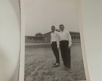 May include: Black and white photograph of two young men standing in front of a school building. They are both wearing white shirts and dark pants. The man on the left is smiling and the man on the right has a serious expression.