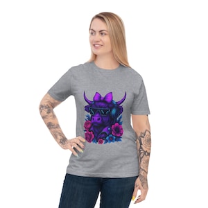May include: A gray t-shirt featuring a purple cartoon cow wearing sunglasses and headphones, adorned with a purple bow and surrounded by pink and blue flowers. The cow graphic is centered on the shirt.