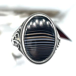 May include: Silver ring with an oval, striped black and gray stone. The ring has a detailed, ornate design on the band and around the stone. The stone has a polished, glossy finish, and the ring is set against a white background.