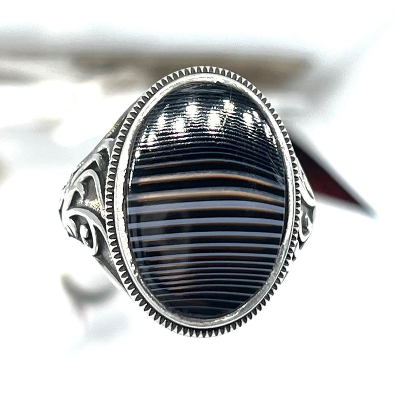 May include: Silver ring with an oval, striped black and gray stone. The ring has a detailed, ornate design on the band and around the stone. The stone has a polished, glossy finish, and the ring is set against a white background.