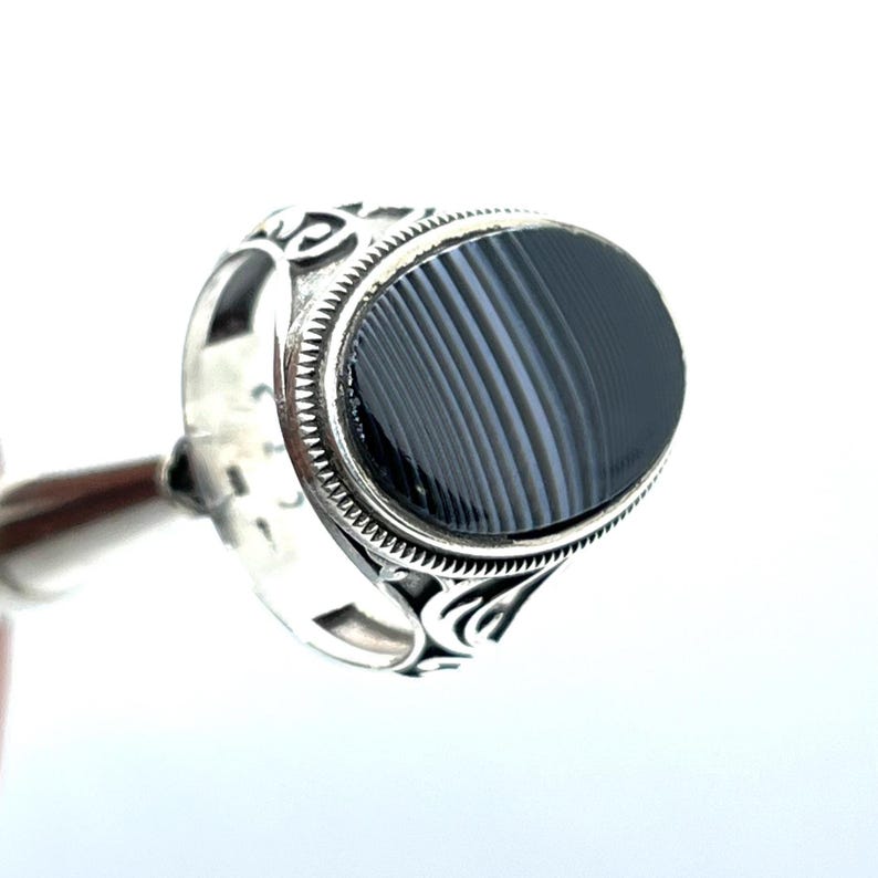 May include: A silver ring with an oval, black stone. The stone has a striped pattern. The ring has decorative scrollwork on the sides and a textured border around the stone. The ring is held up by a silver rod.