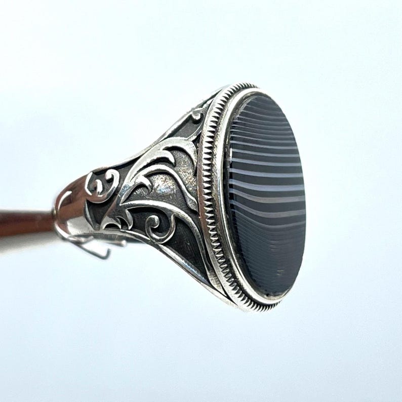 May include: A silver ring with an oval, black and gray striped stone. The ring has an ornate, detailed design on the band with a textured border around the stone. The ring is held up by a wooden stick.
