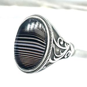 May include: A silver ring with an oval-shaped, black and white striped stone. The ring has an ornate, engraved design on the band and around the stone. The stone's pattern creates a layered effect.