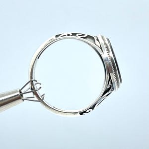 May include: A silver ring with an oval, dark-colored stone. The ring has intricate filigree detailing on the band and around the stone setting. The ring is held by a metal tool, showcasing the design and craftsmanship.