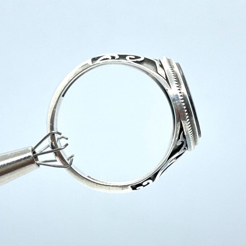 May include: A silver ring with an oval, dark-colored stone. The ring has intricate filigree detailing on the band and around the stone setting. The ring is held by a metal tool, showcasing the design and craftsmanship.