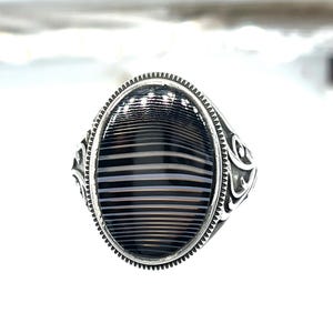 May include: A silver ring with an oval-shaped, striped black and white stone. The ring has a detailed, patterned design around the stone and on the band. The stone's stripes are horizontal, creating a bold visual effect.