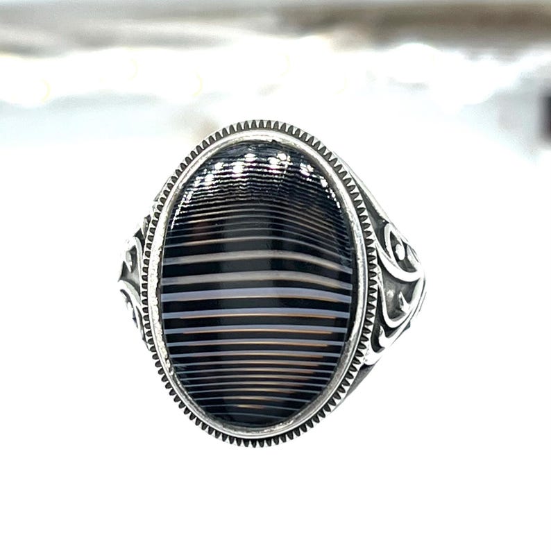 May include: A silver ring with an oval-shaped, striped black and white stone. The ring has a detailed, patterned design around the stone and on the band. The stone's stripes are horizontal, creating a bold visual effect.