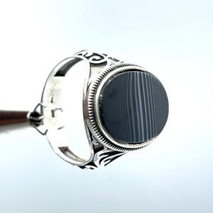 May include: A silver ring with an oval, black and gray striped stone. The ring has an ornate design on the band and around the stone. The ring is displayed against a white background.