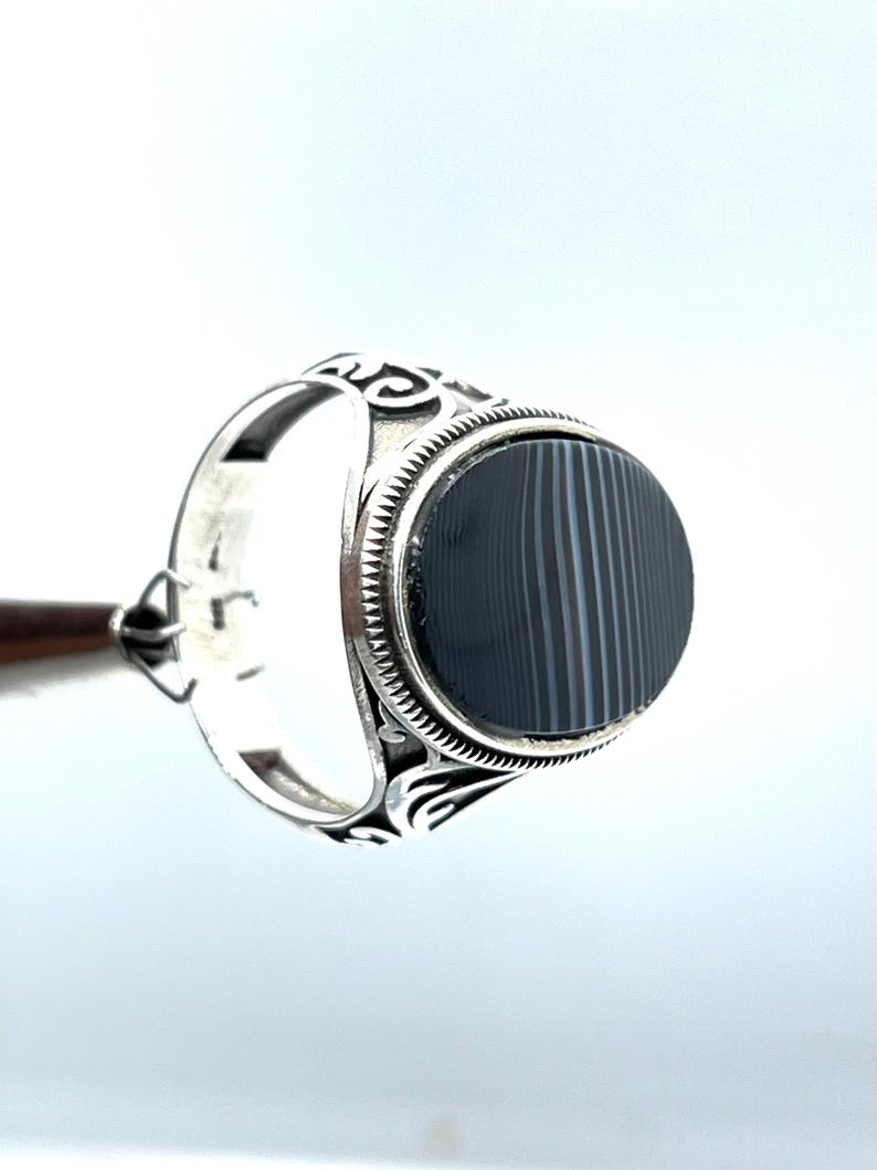 May include: A silver ring with an oval, black and gray striped stone. The ring has an ornate design on the band and around the stone. The ring is displayed against a white background.