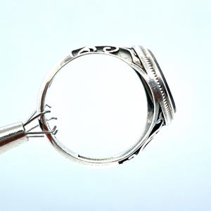 May include: Close-up of a silver ring with an oval, dark-colored stone. The ring has an ornate, filigree design on the band and around the stone setting. The ring is held by a jewelry tool, showcasing the intricate details and craftsmanship.