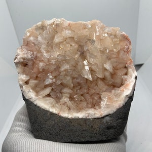 Könnte beinhalten: Ein Geode mit einem rosa und weißen Inneren, gefüllt mit kleinen, klaren Kristallen. Der Geode ist teilweise geöffnet und zeigt die Kristalle im Inneren. Die Außenseite des Geoden ist dunkelgrau.