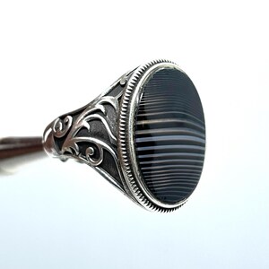 May include: A silver ring with an oval, black and gray striped stone. The ring has ornate, swirling designs on the band and around the stone setting. The stone is approximately 1 inch long.